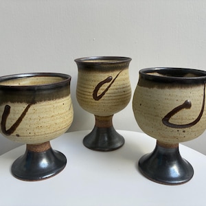 May include: Three ceramic goblets featuring a speckled cream-colored body with a dark brown rim and a solid dark brown pedestal base. Each goblet displays a single dark brown hand-painted curved line accent on the exterior surface of the bowl.