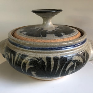 May include: A ceramic casserole dish with a lid. The dish is decorated with a black and white floral pattern and has a blue rim. The lid has a knob on top.