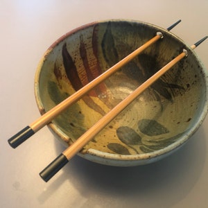 May include: A ceramic bowl with a floral design in shades of green, brown, and yellow, paired with two wooden chopsticks. The chopsticks have black tips and are resting inside the bowl.