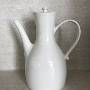 May include: A white ceramic coffee pot with a curved handle and a spout. The pot has a lid with a small knob. The design is simple and elegant, suitable for serving coffee or tea. The pot is approximately 10 inches tall.