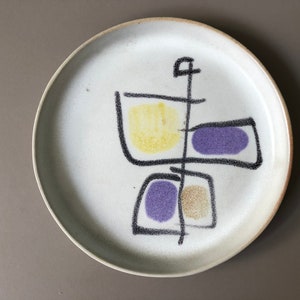May include: A white ceramic plate with a black and white abstract design featuring yellow, purple, and brown accents.