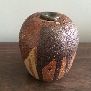 May include: A small, round, brown and tan ceramic vase with a textured surface. The vase has a small opening at the top.