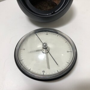 May include: A round, white-faced clock with black numerals and hands, encased in a clear, domed cover. The clock sits on a white surface, with a black, ribbed container in the background.