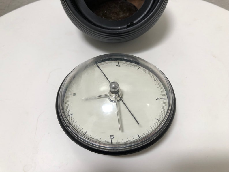 May include: A round, white-faced clock with black numerals and hands, encased in a clear, domed cover. The clock sits on a white surface, with a black, ribbed container in the background.