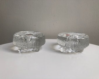 May include: Two clear glass candle holders with a textured surface and three legs each. The candle holders are on a white surface.