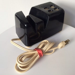 May include: A black electric knife sharpener with a white cord and plug. The sharpener has two slots for sharpening knives.