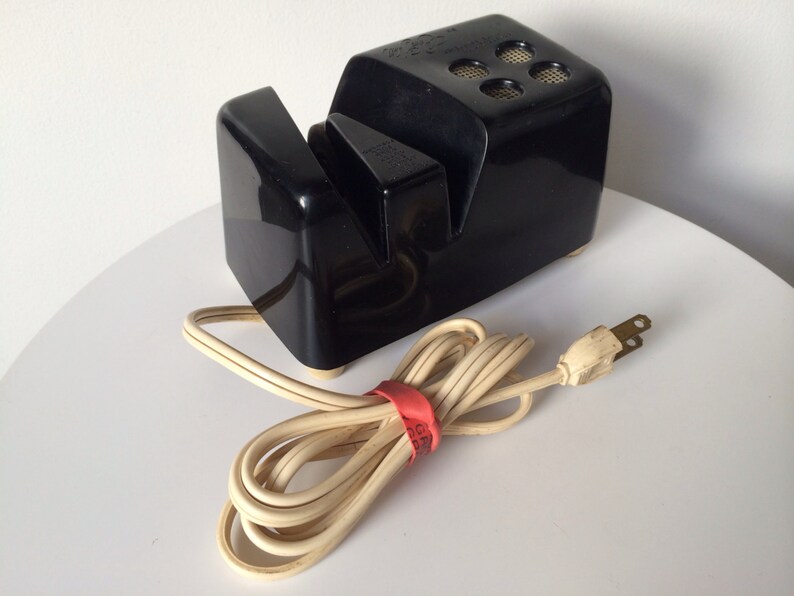 May include: A black electric knife sharpener with a white cord and plug. The sharpener has two slots for sharpening knives.