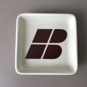 May include: Square, off-white ceramic dish with a brown graphic design. The design is a stylized letter "B" composed of four connected shapes. The dish has rounded corners and a slightly raised edge.