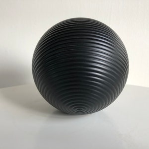 May include: A black, spherical decorative object with a textured, ridged surface. The ridges spiral around the sphere, creating a pattern. The object is likely made of a solid material and is designed for display.