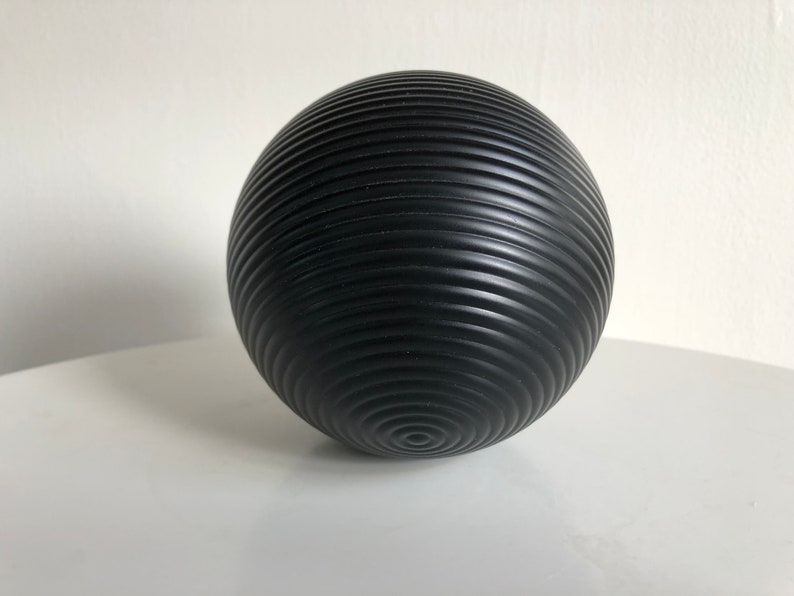 May include: A black, spherical decorative object with a textured, ridged surface. The ridges spiral around the sphere, creating a pattern. The object is likely made of a solid material and is designed for display.