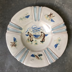 May include: A white ceramic plate with a blue and brown design of two figures pulling a net from the water. The plate has a scalloped edge and a blue and white glaze.