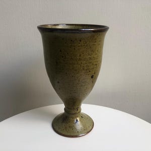 May include: A handmade, speckled ceramic goblet with a wide rim and a pedestal base. The goblet has a muted olive green color with darker brown accents around the rim and base. The piece is likely for decorative use.