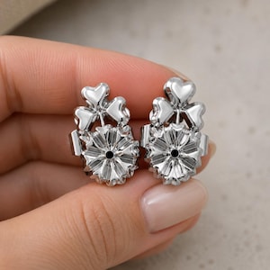 Può includere: Una coppia di sollevatori per orecchini color argento con un design decorativo a trifoglio a tre foglie sulla parte superiore. Ogni sollevatore ha una base circolare smerlata con un foro centrale per sostenere gli orecchini.