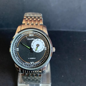May include: A silver-toned ZentRa wristwatch with a black face, featuring white and green accents. The watch has a metal band and displays the text "17 JEWELS" and "MADE IN GERMANY". The watch is displayed on a clear stand.