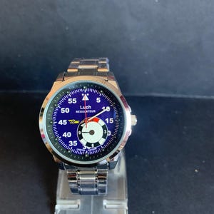 May include: A silver wristwatch with a blue face and white and red accents. The watch face displays the text "Luch REGULATEUR LIMITED EDITION" and has a silver band. The watch is displayed on a clear plastic stand.