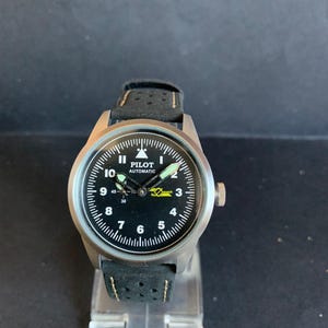 Op de afbeelding: Een zilverkleurig Pilot Automatic horloge met een zwarte wijzerplaat en witte cijfers. Het horloge heeft een zwarte leren band met perforaties. De wijzerplaat toont de tekst "PILOT AUTOMATIC".