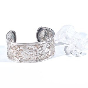 May include: A silver cuff bracelet with a floral design. The bracelet is made of metal and has a hammered finish.