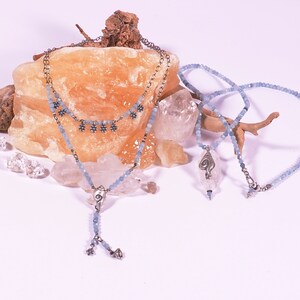 May include: Two delicate necklaces with light blue beads and silver accents. One necklace features a pendant with a silver swirl design. The necklaces are displayed on a light orange rock and a piece of driftwood.
