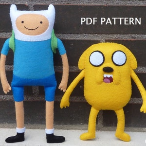 Puede incluir: Juguetes de peluche de fieltro de Finn y Jake de la serie de dibujos animados Adventure Time. Finn lleva una camisa azul y pantalones cortos azules. Jake es amarillo con una nariz negra y ojos blancos. El texto "PDF PATTERN" está impreso en una pared de ladrillo detrás de los juguetes.