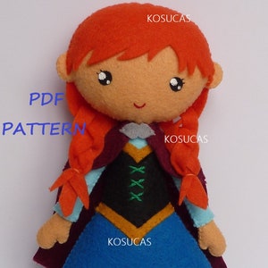 Puede incluir: Muñeca de fieltro hecha a mano con cabello rojo trenzado, vestida con un vestido azul, un corpiño negro y una capa burdeos. La muñeca tiene una cara sonriente y está etiquetada con el texto "PDF PATTERN" y la marca "KOSUCAS".