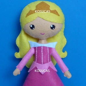 May include: A pink felt doll of a princess with blonde hair and a crown. The doll is wearing a pink dress with a white collar and has a stitched design on the bodice. The doll is holding its hands out in front of it. The text "PDF PATTERN" is written in white letters on a black background at the bottom of the image.