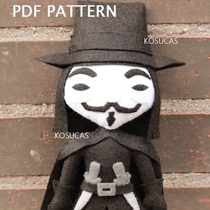 May include: A felt plush toy of the character V for Vendetta. The toy is wearing a black hat, a black coat, and a white mask with a black mustache and goatee. The toy is standing in front of a brick wall.