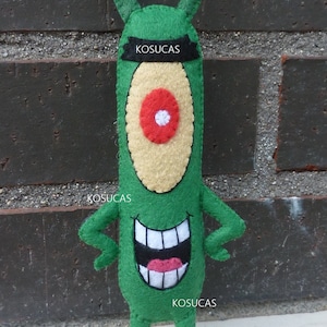 May include: Green felt plush toy with a large eye, a toothy grin, and the text "KOSUCAS" on the headband, chest, and feet.