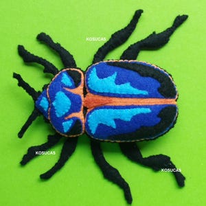 Könnte beinhalten: Ein Filzinsekt mit blauem und orangefarbenem Design auf grünem Hintergrund. Der Käfer hat schwarze Beine und Fühler. Der Text "PDF PATTERN" befindet sich oben im Bild.