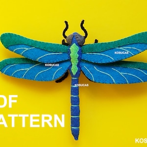 Op de afbeelding: Een handgemaakte vilten libel in blauw- en groentinten, met witte stikseldetails. De libel heeft een gesegmenteerd lichaam en gebogen antennes. De tekst "PDF PATTERN" wordt in witte letters op een gele achtergrond weergegeven. De merknaam "KOSUCAS" is ook zichtbaar.