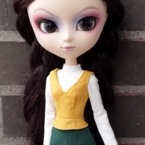 May include: A doll with long black hair, wearing a white long-sleeved shirt, a yellow vest, and a green skirt. The doll has large eyes with pink and blue eyeshadow and is standing in front of a brick wall. The text "KOSUCAS" is visible.