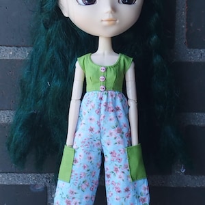 May include: A doll with long green hair wearing a green top with pink buttons and a blue floral jumpsuit with green accents. The doll has large eyes and is posed in front of a brick wall. The text "KOSUCAS" is visible at the bottom right.