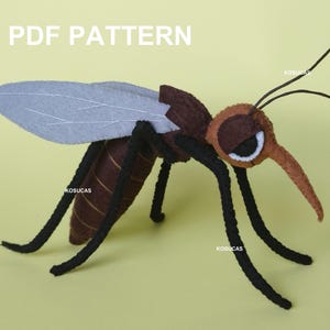 Op de afbeelding: Een handgemaakt vilten muggenmodel met bruine, zwarte en grijze details. Het insect heeft grote vleugels, lange poten en een lange steeksnuit. De tekst "PDF PATTERN" staat bovenaan. De mug staat op een geelgroene achtergrond.