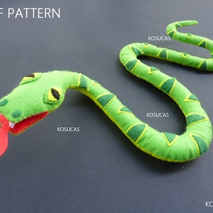 Pode incluir: Uma cobra de feltro verde com língua vermelha e olhos amarelos. A cobra tem um padrão verde e amarelo. A imagem inclui o texto "PDF PATTERN" e "KOSUCAS".