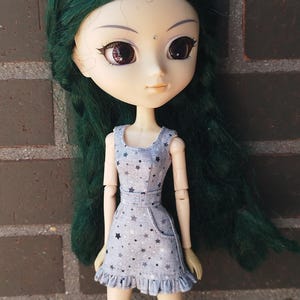 May include: A doll with green hair and large eyes, wearing a light blue dress with a star pattern. The dress has a ruffled hem and a small pocket. The doll is posed in front of a brick wall.