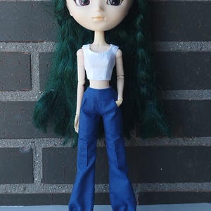 May include: A doll with green hair wearing a white crop top and blue cargo pants. The doll has large eyes and is standing in front of a gray brick wall. The pants have pockets on the sides. The doll's name is Kosucas.