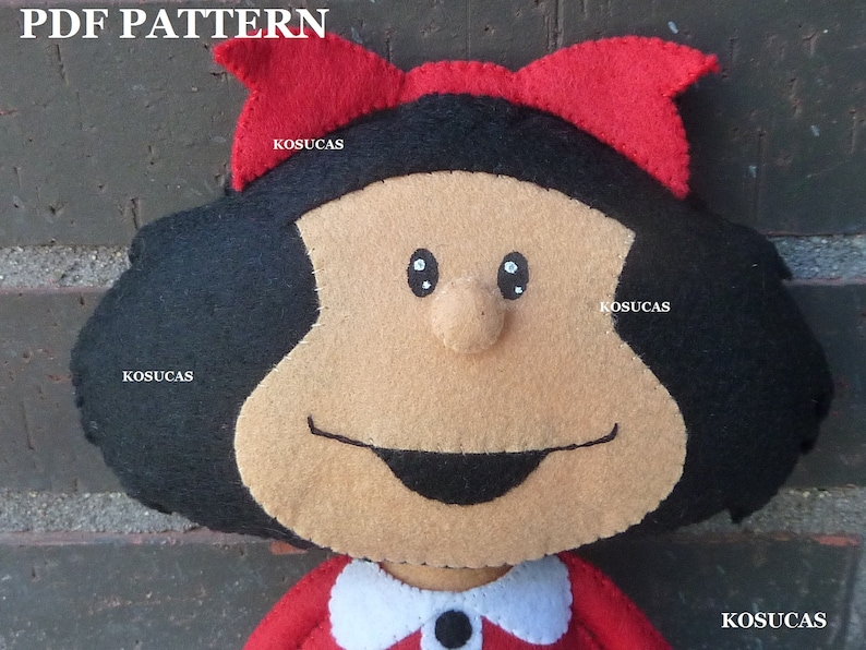 May include: A felt doll with black hair, a red bow, and a smiling face. The doll is wearing a red dress with a white collar and a black button.