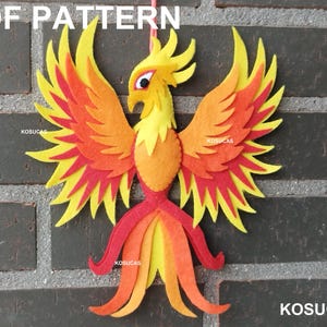 Puede incluir: Decoración de fieltro de un fénix en tonos amarillos, naranjas y rojos, con el texto "PDF PATTERN" y "KOSUCAS". El ave tiene las alas extendidas y una cola detallada, colgada sobre un fondo de ladrillos.