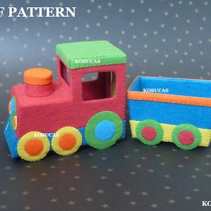 Könnte beinhalten: Ein Spielzeugzug aus Filz mit einer roten Lokomotive, einem blauen Güterwagen und bunten Rädern. Der Zug hat ein grünes Dach und einen kleinen orangenen Schornstein. Die Worte "KOSUCAS" sind auf der Lokomotive und dem Güterwagen aufgedruckt.