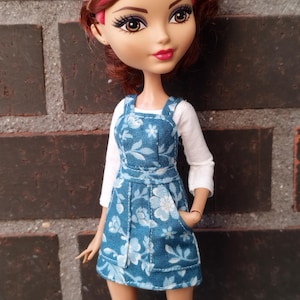 Peut inclure: Une poupée aux cheveux roses et rouges, vêtue d'une chemise blanche à manches longues et d'une robe bleue à imprimé floral. La poupée a les yeux marron et se tient devant un mur de briques. Le texte "KOSUCAS" est visible en bas.