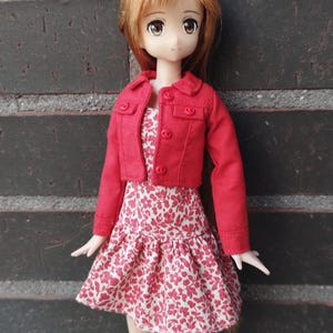 Puede incluir: Una muñeca vestida con una chaqueta roja y un vestido con estampado floral. La chaqueta tiene tres botones y un cuello. El vestido es blanco con un estampado floral rojo. La muñeca tiene ojos marrones y cabello castaño claro. El texto "KOSUCAS" es visible.