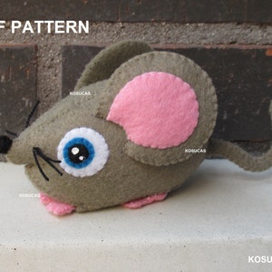 Könnte beinhalten: Eine graue Filzmaus mit rosa Ohren und einem blauen Auge. Die Maus hat einen langen, grauen Schwanz. Die Maus ist ein handgemachtes Spielzeug.