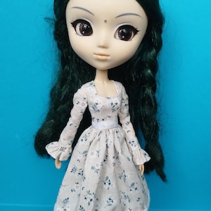May include: A doll with dark green hair and large, expressive eyes, wearing a floral print dress with long sleeves and a white belt. The dress has a scalloped hem. The doll is set against a bright blue background. The text "KOSUCAS" is visible.
