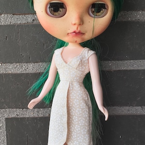 May include: A doll with long green hair and large brown eyes wearing a cream-colored sleeveless dress with a small white floral pattern. The dress features a V-neckline and a wrap-style skirt. The word KOSUCAS is printed in the bottom right corner.