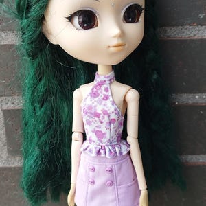 May include: A doll with green hair and large eyes, wearing a halter top with a floral pattern and a lavender skirt with buttons. The doll's outfit is detailed, and the doll is posed in front of a brick wall. The text "KOSUCAS" is visible.