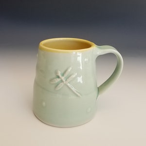 May include: A light blue ceramic mug with a yellow rim and a dragonfly design. The mug has a rounded shape and a handle.