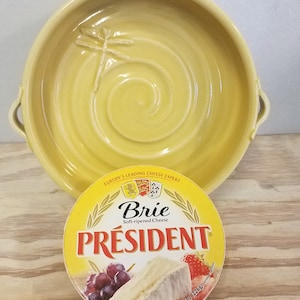 May include: A yellow ceramic dish with a dragonfly design and a round brie cheese wheel with the label "President Brie Soft-ripened Cheese" and a net weight of 8 ounces (226 grams).
