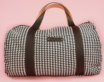 Puede incluir: Una bolsa de viaje de lona con estampado de pata de gallo en blanco y negro con asas y correas de cuero marrón. La bolsa tiene cierre de cremallera y una etiqueta de cuero con la palabra "CRAFT" en relieve.