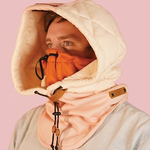 May include: A white hooded neck gaiter with a quilted design and a brown leather patch. The gaiter has a peach-colored fleece lining and a black drawstring. The gaiter is worn by a person with a peach-colored fleece face mask.
