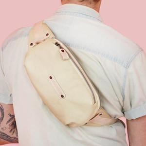 Puede incluir: Una riñonera de cuero beige claro con cierre de cremallera y correa ajustable. La bolsa se lleva en la espalda de una persona que lleva una camisa azul claro.