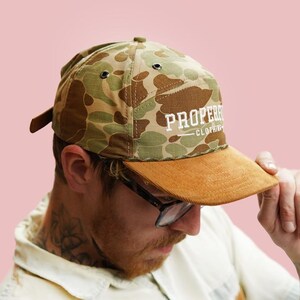 Puede incluir: Gorra de b&eacute;isbol con estampado de camuflaje y visera de ante marr&oacute;n. La gorra tiene el texto "PROPER CLOTHING" en letras blancas. El fondo es rosa claro liso.
